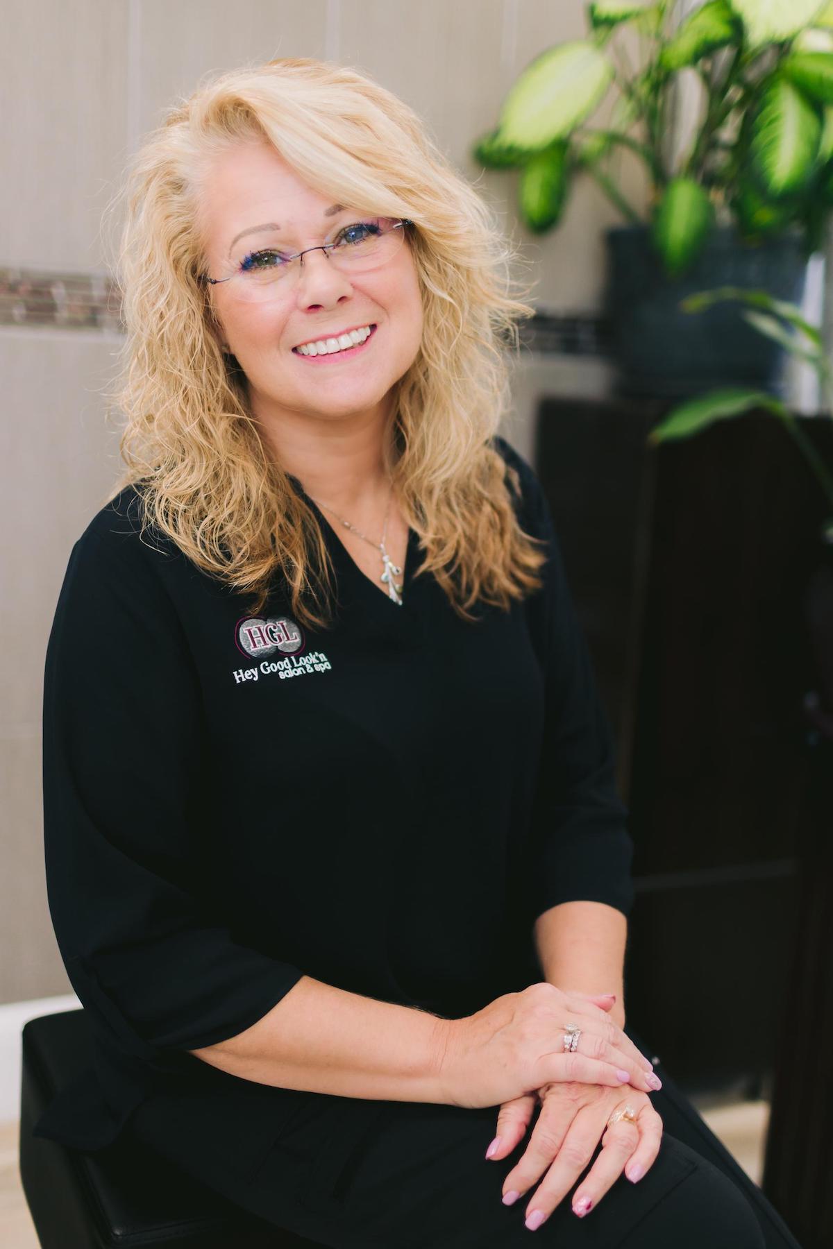 a professional blonde woman with glasses sits on a chair posing for a personal branding photoshoot, representing her brand as a skilled hair and makeup artist.