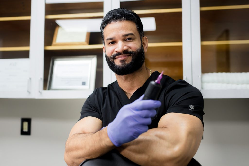 a bearded man wearing purple gloves holds a professional tool, highlighting his personal branding as a skilled Scalp Micropigmentation technician.