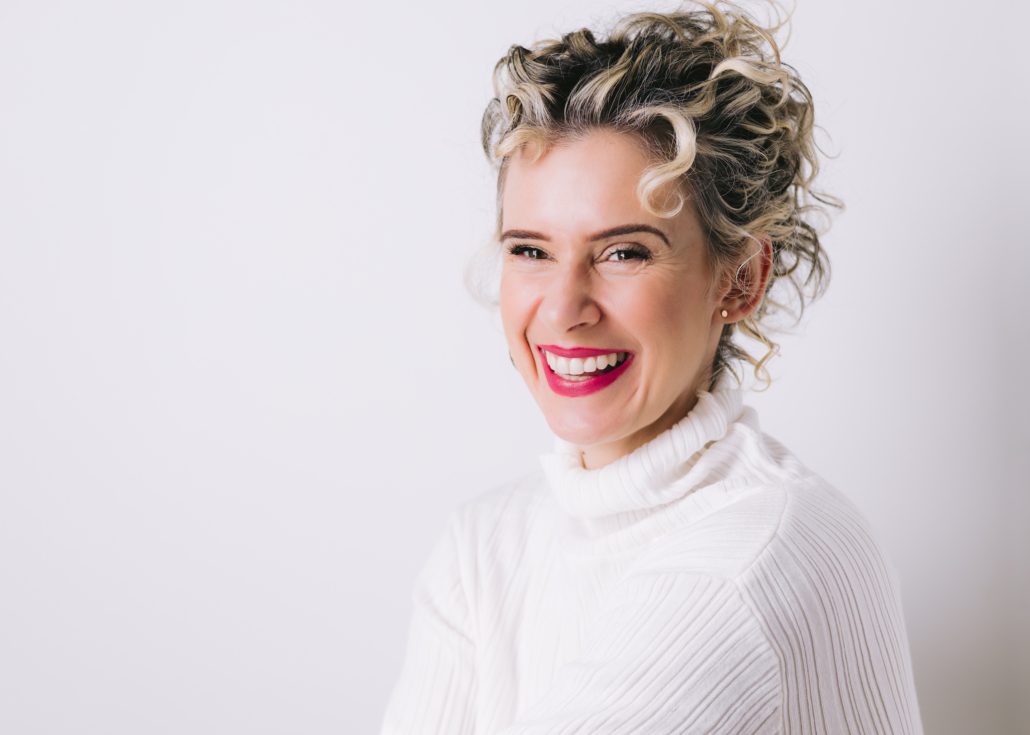 A curly hair female in a white sweater smiling in front of a simple white backdrop.