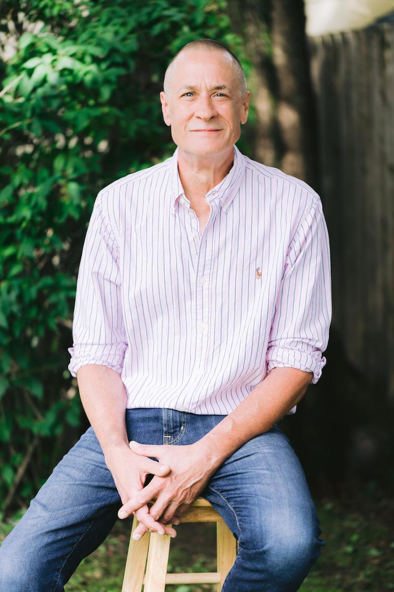 A mature man in a pink stripe shirt and jeans sitting on a stool in a backyard posing for headshot.