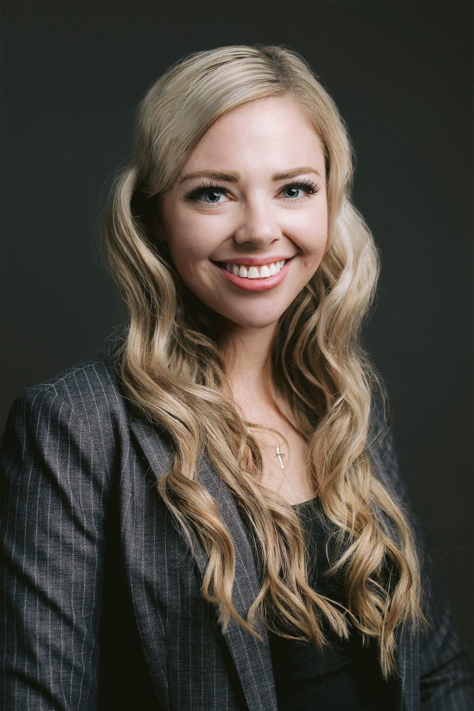 a confident blonde professional poses in a business suit, smiling for a headshot with a dark background, captured during a branding session.